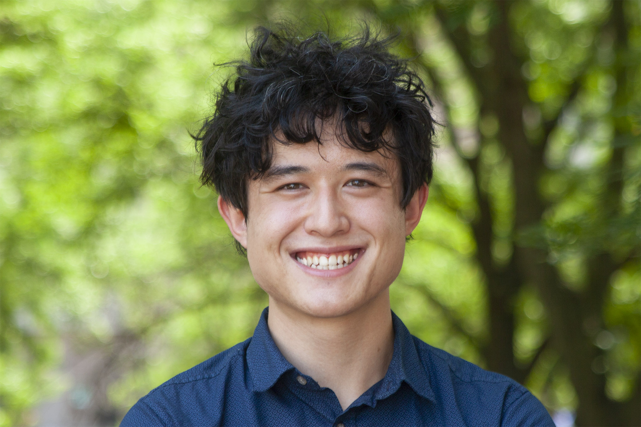 Person in a blue shirt with black curly hair standing in front of a background of green trees.