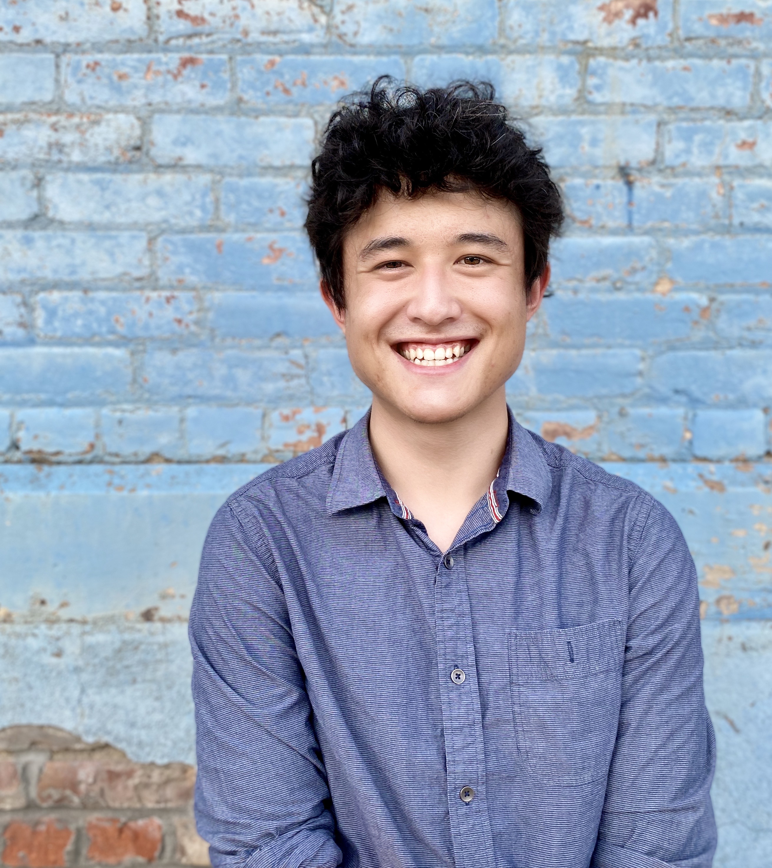 Person in a blue shirt with black curly hair standing in front of a background of a light blue brick wall.